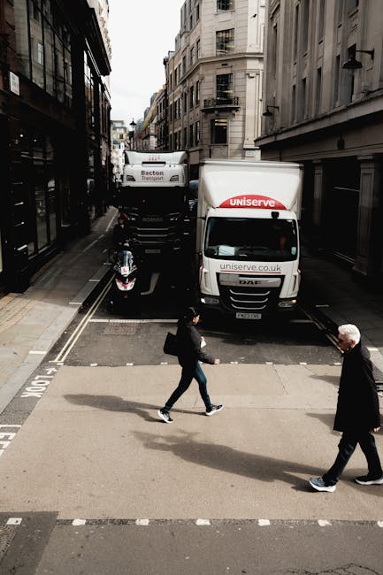 The image shows a narrow urban street with tall, multi-storey buildings on either side, featuring facades with balconies and large windows. Two white moving trucks branded with 'Uniserve' and 'Becton