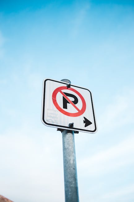 A rectangular illuminated sign affixed to a light-colored brick wall indicates that parking is not allowed in the alley, featuring a red circle with a diagonal line crossing out a car symbol and the t