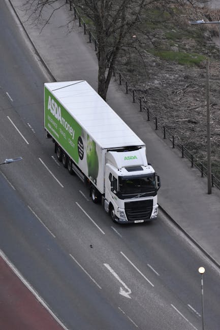 An aerial view of a white delivery truck with the ASDA Money logo parked on a paved urban street. The truck is positioned parallel to the sidewalk, with its cab facing forward and the cargo area exten