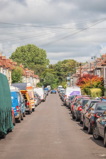 The image depicts a residential street in Kennington during spring, with a clear view down the road lined on both sides by parked cars, including a mix of sedans and hatchbacks. The street is bordered