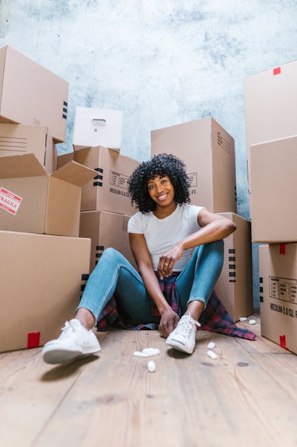 A man and a woman are standing inside a doorway, each holding cardboard boxes containing household items during a home relocation process. The man is wearing a maroon shirt and loose orange trousers, 